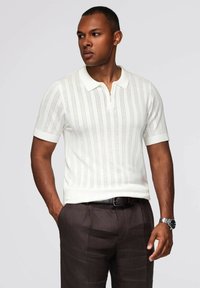 Polo de punto blanco de manga corta con patrones de rayas verticales y cuello con cremallera, combinado con pantalones marrón oscuro.