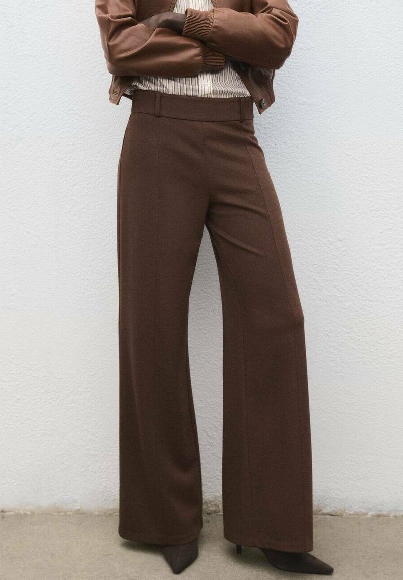 Personne debout, bras croisés, portant un pantalon large marron, une veste assortie et des talons aiguilles pointus, devant un mur clair.