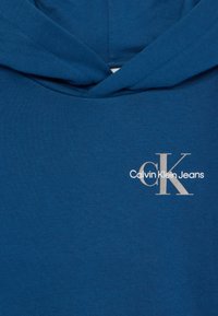 Modrá bavlnená mikina s kapucňou, prednou vreckom a šnúrkami na kapucni, s bielym logom, na ktorom je elegantným písmom napísané "Calvin Klein Jeans".