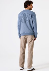 Sudadera azul con un logo circular texturizado en la parte de atrás, combinada con pantalones de pana color beige y zapatillas blancas con rayas negras.