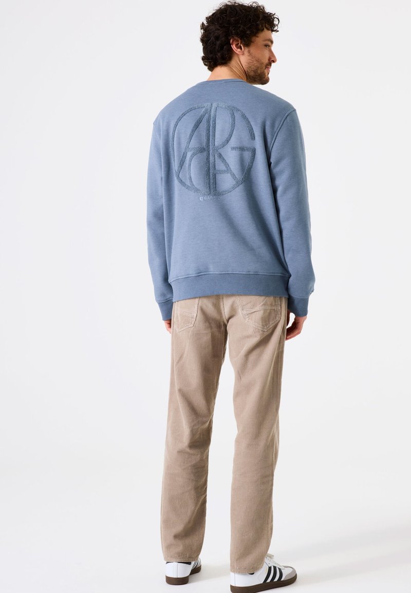 Sudadera azul con un logo circular texturizado en la parte de atrás, combinada con pantalones de pana color beige y zapatillas blancas con rayas negras.