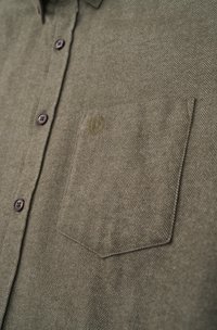 Olive green button-up shirt featuring a herringbone pattern, with a left chest pocket and dark brown buttons. Small embroidered logo on pocket.