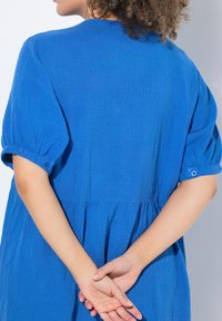 Blusa blu, con texture, maniche corte a sbuffo e vita arricciata. Presenta dettagli con bottoni sulle maniche e una silhouette ampia.