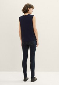 Mujer con cabello corto y rizado de espaldas, vistiendo un top de punto sin mangas de color azul marino, jeans oscuros ajustados y zapatos de cuero negros.