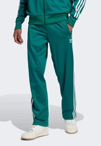 Muškarac nosi tirkizne Adidas trenirke s bijelim prugama uz strane, bijele tenisice i odgovarajuću jaknu s rukom u džepu.