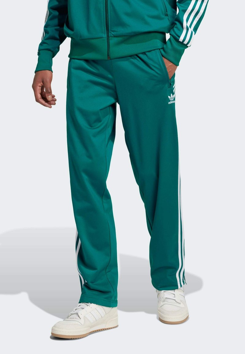 Muškarac nosi tirkizne Adidas trenirke s bijelim prugama uz strane, bijele tenisice i odgovarajuću jaknu s rukom u džepu.