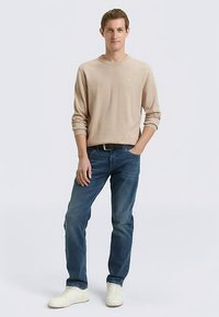 Pull beige à manches longues, jean en denim bleu, baskets blanches, coupe ample, texture unie, sans motifs ni accents visibles. Position debout.