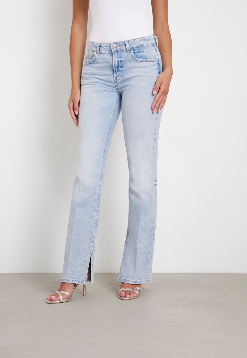 Jeans acampanados de color azul claro con cortes frontales, hechos de denim. Cuenta con un diseño clásico de cinco bolsillos y cierre de botón. Combinados con tacones plateados con tiras.