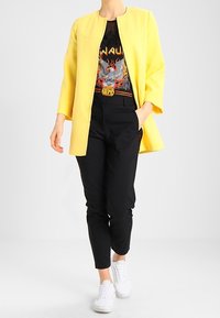 Abrigo amarillo hasta la rodilla con cuello redondo, llevado sobre una camiseta gráfica negra. Un pantalón negro a medida y zapatillas blancas completan el conjunto.