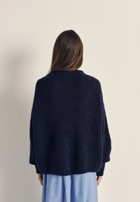 Navy blue knitted sweater with a loose fit, textured fabric, wide sleeves, and a rounded neckline. Worn over a light blue striped skirt.