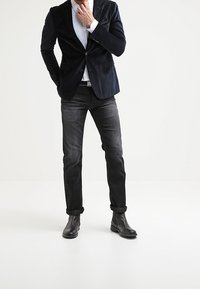 Blazer di velluto blu navy sopra una camicia a righe bianche e blu, abbinato a jeans scuri e stivali neri alla caviglia, creando un aspetto coeso e raffinato.