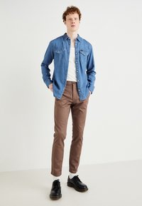 Denim shirt in blue with two chest pockets, paired with brown trousers and black leather shoes. Background is neutral and minimalistic.