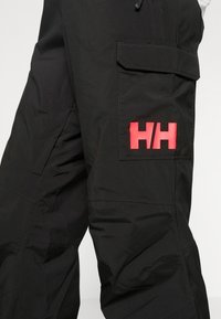 Černé cargo kalhoty vyrobené z odolného materiálu, s velkou kapsou na boku a výrazným červeným logem. Hladká textura s minimalistickým designem.