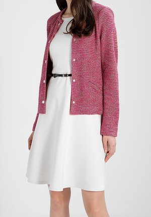 Kobieta ubrana w białą sukienkę bez rękawów z czarnym paskiem i teksturowany różowy sweter zapinany na guziki na wierzchu.