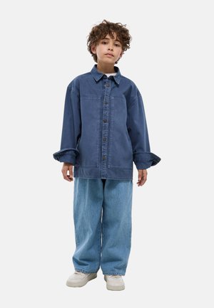 Enfant aux cheveux bouclés portant une veste en jean bleue surdimensionnée, un jean large bleu clair et des baskets blanches, debout devant un fond uni.