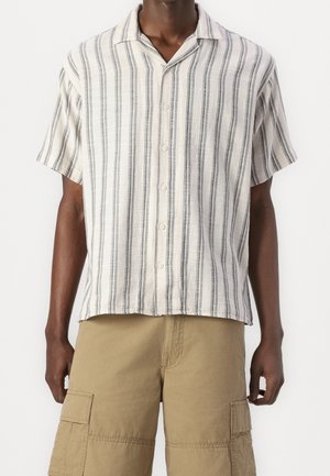 Homme portant une chemise beige à manches courtes, boutonnée, avec des rayures noires verticales, et un short cargo beige jusqu'aux genoux.