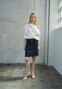 Witte off-shoulder blouse met verticale strepen, gecombineerd met een donkere denim rok en nude hakken, tegen een neutrale achtergrond.