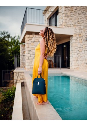 Gelbes plissiertes Jumpsuit, dunkelgrüne Tasche mit einem silbernen Reißverschluss, barfuß am Rand eines Pools stehend, neben einem Steingebäude und Wasser.