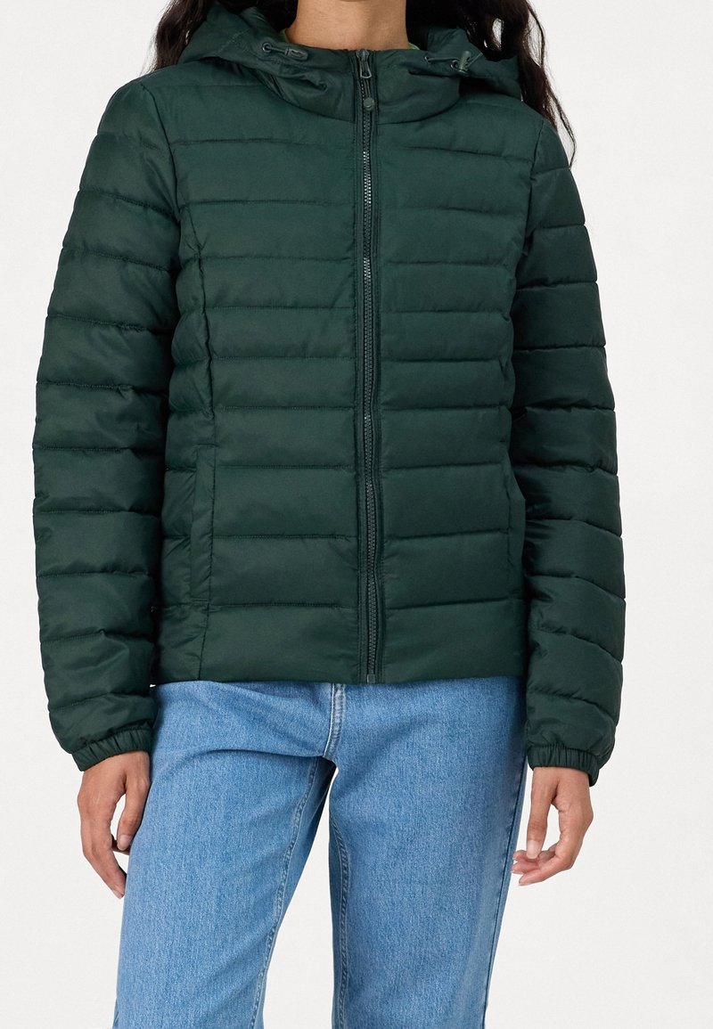 Polstret grøn jakke med lynlås foran, hætte, elastiske manchetter og lodret quilted design. Kombination med lyseblå jeans. Glat, skinnende tekstur.