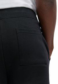 Zwarte joggers met een geribbelde tailleband hebben een vierkant achterzakje. De stof lijkt zacht met een gladde textuur en toont een minimalistisch ontwerp.