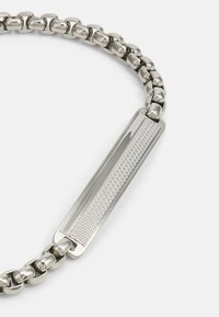 Collar de cadena de metal plateado con un cierre rectangular texturizado con un patrón de diamantes, y eslabones pulidos para un acabado suave.