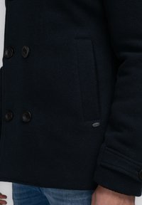 Abrigo de lana azul marino oscuro con cierre de botones de doble botonadura, dos bolsillos laterales y una etiqueta de metal de la marca en el lado izquierdo.