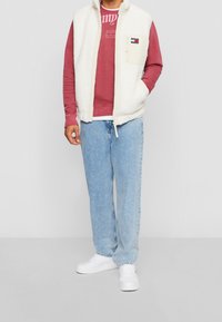 Biały polarowy bezrękawnik na bordowej bluzie z kapturem, jasnoniebieskie dżinsy i białe trampki; posiada kieszeń na piersi i zapięcie na zamek.