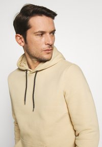 Beige hoodie gemaakt van zachte stof, met een voorzak, zwarte trekkoorden met witte accenten, en een ontspannen pasvorm.