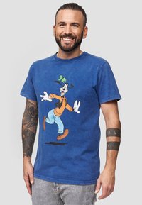 Sinisiä puuvillapaitoja, joissa on Goofy-hahmon grafiikka oranssissa ja sinisessä asussa. Lyhyet hihat, pyöreä kaula-aukko, rento malli ja pehmeä tekstuuri.