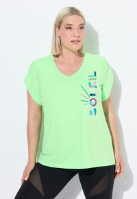 Lys grøn trænings t-shirt, v-hals design, korte rullede ærmer, har flerfarvet grafisk tekst på siden, parret med sorte tights.