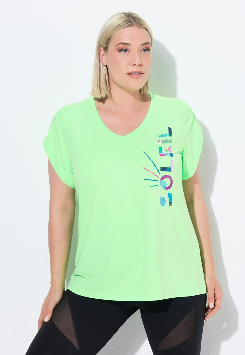 Lys grøn trænings t-shirt, v-hals design, korte rullede ærmer, har flerfarvet grafisk tekst på siden, parret med sorte tights.