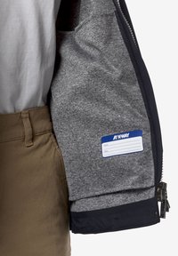 Η διαφημιστική μπλούζα K-Way έχει γκρι fleece εσωτερικό, μπλε ταμπελάκι, ναυτική επένδυση και λεπτομέρεια με τσέπη με φερμουάρ. Συνδυάζεται με καμηλό παντελόνι.