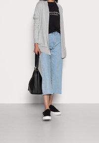 Žena v světle šedém kardiganu, černém tričku s logem, světle modrých širokých zkrácených jeansách, černých teniskách a držící černou kabelku.