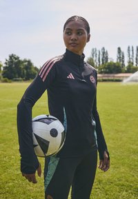 Svart treningjakke med rosa striper, glidelås krage, og turkise aksenter. Svarts bukser. Spiller holder en svart og hvit fotball på gressbane.