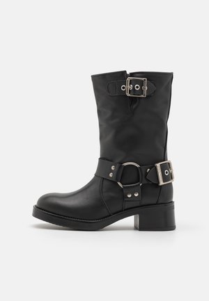 Bottine en cuir noir avec un bout arrondi, un talon épais et une sangle ajustable avec une boucle en argent et un détail en anneau. Texture lisse.