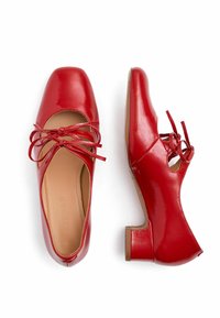 Zapatos de charol rojos con punta cuadrada, tacón bloque de altura media y cordones decorativos de cinta. Textura suave y detalles recortados.