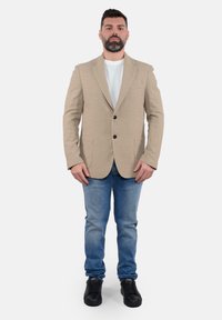 Blazer beige testurizzato con due bottoni neri, abbinato a una camicia bianca, jeans blu e sneakers nere.