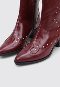 Bottines en cuir bordeaux avec des pointes pointues, ornées de clous dorés décoratifs et d'une fermeture éclair sur le côté. Les talons carrés améliorent la stabilité.
