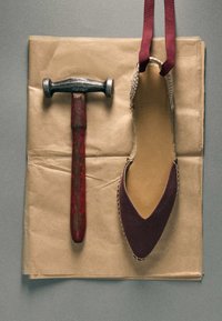 Marteau rustique avec manche rouge usé à côté d'une chaussure plate à bout pointu avec semelle en jute tressée et lanières bordeaux sur papier kraft.