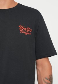 Musta t-paita puuvillasta, jossa on punainen graafinen teksti "Walt's Waffles" vasemmassa rintataskussa. Lyhyet hihat, normaali istuvuus.
