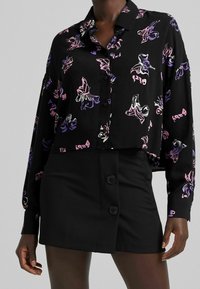 Black cropped button-up shirt with colorful butterfly patterns, paired with a black skirt featuring two dark buttons at the front.