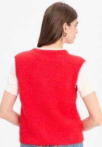 Rote Strickweste mit strukturiertem Finish, ärmelloses Design und runder Ausschnitt. Über einem weißen kurzärmligen Hemd getragen.