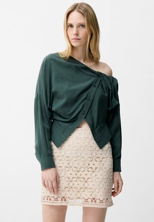 Femme portant un chemisier asymétrique boutonné vert foncé et une jupe beige en crochet, debout devant un fond clair uni.