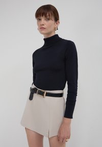 Femme portant un pull à col roulé noir, une jupe beige avec une fente latérale, une ceinture noire, des boucles d'oreilles en or et des bagues en or sur fond uni.