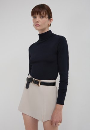 Frau trägt einen schwarzen Rollkragenpullover, einen beigen Rock mit seitlichem Schlitz, einen schwarzen Gürtel, goldene Creolen und goldene Ringe vor einem schlichten Hintergrund.
