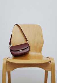 Chaise en bois aux bords arrondis portant un sac à bandoulière en cuir marron foncé suspendu au dossier, devant un mur uni.