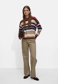 Maglione a righe con colori rosso, giallo, blu navy e beige, abbinato a pantaloni beige a gamba larga e ballerine marrone scuro. Borsa con frange molto evidente.