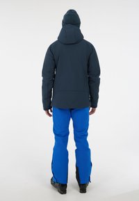 Skijack in donkerblauw met capuchon, gecombineerd met felblauwe skibroeken. Zwarte skischoenen zichtbaar, met ritssluitingen op de broekspijpen.