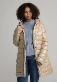 Beige pufferjack met gestikt ontwerp, capuchon en rits aan de voorkant. Gepaarde met een lichtblauwe geribbelde trui en blauwe jeans. Gladde textuur.