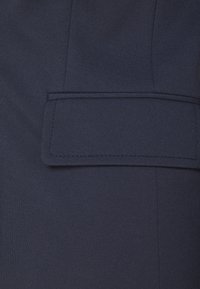 Gros plan d'une poche en tissu bleu marine avec rabat et coutures visibles sur un vêtement.